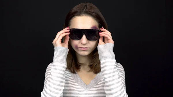 Una mujer joven se pone gafas oscuras para cerrar moretones en la cara. Una mujer con moretones en la cara. Pelea en una familia joven. Violencia doméstica. Sobre un fondo negro — Foto de Stock