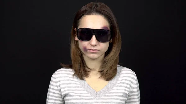 O tânără poartă ochelari de soare pentru a-şi închide vânătăile pe faţă. O femeie cu vânătăi pe faţă. Ceartă într-o familie tânără. Violenţa domestică. Pe un fundal negru — Fotografie, imagine de stoc