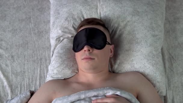 若い男は睡眠マスクで眠りに落ちることはできません。男は自分の部屋のベッドに横たわって不眠症に苦しんでいます。上からの眺め. — ストック動画