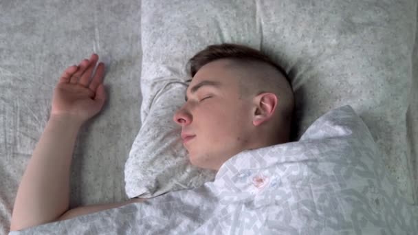 Um jovem está a dormir debaixo dos lençóis. Um homem está deitado na cama no quarto. Vista de cima . — Vídeo de Stock