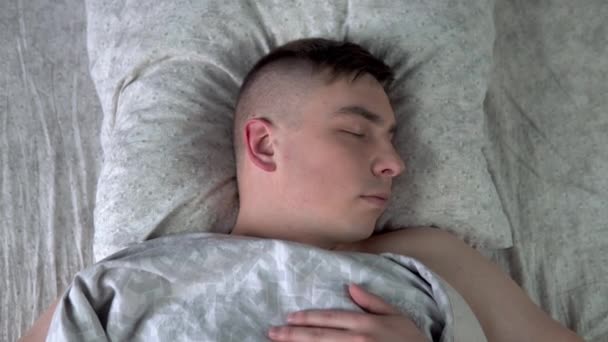 Un joven está durmiendo bajo las sábanas. Un hombre yace en la cama de su habitación. Vista desde arriba . — Vídeos de Stock