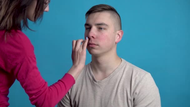 Макияж наносят на молодого человека. Девушка наносит макияж мужчине перед съемкой на синем фоне . — стоковое видео