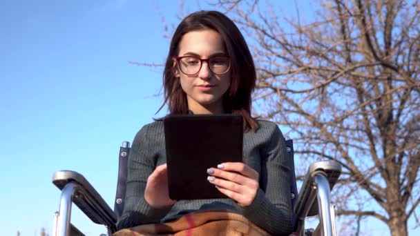 Un joven en silla de ruedas con una tableta. El hombre discapacitado en la naturaleza está en correspondencia en una tableta . — Vídeos de Stock