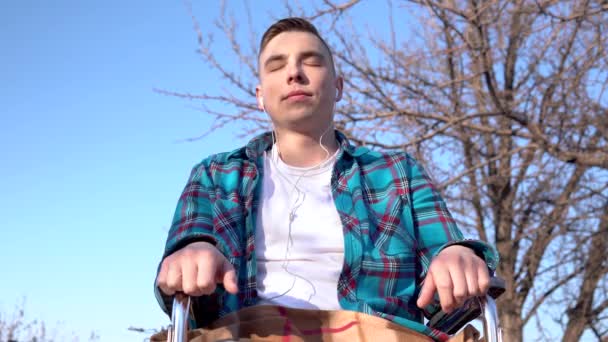 Młody człowiek na wózku inwalidzkim ze słuchawkami. Niepełnosprawny człowiek w naturze ze słuchawkami w uszach. — Wideo stockowe