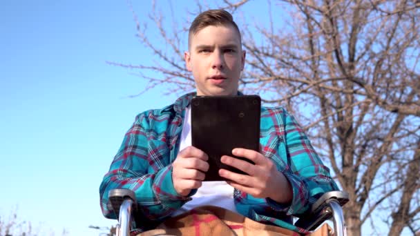Jovem numa cadeira de rodas com um tablet. Homem com deficiência na natureza está em correspondência em um tablet . — Vídeo de Stock