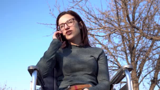 A young woman on a wheelchair is talking on the phone. Disabled girl in nature with a smartphone. — Stock Video