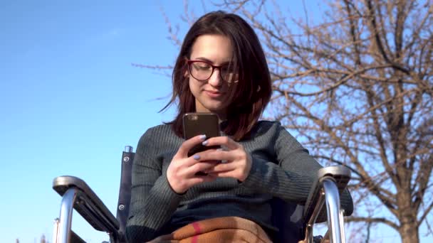 Junge Frau im Rollstuhl mit Handy. Behinderte Mädchen in der Natur kommunizieren auf dem Smartphone. — Stockvideo