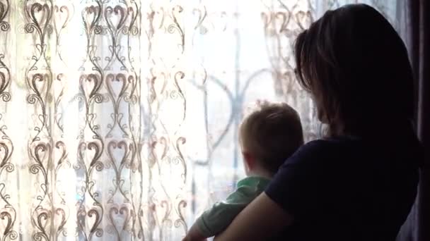 Młoda matka trzyma dziecko w ramionach. Kobieta z dzieckiem patrząca przez okno odsuwając zasłonę.. — Wideo stockowe