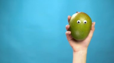 Mango bir kadının elinde gözleri var. Mango biberi çerçevenin içine atlar ve mavi bir arkaplanda etrafına bakar. Kadın eli yakın çekim.