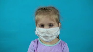 Mavi arka planda tıbbi maskeli küçük bir kız. Kız başparmak gösteriyor..