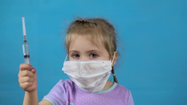 Tıbbi maskeli küçük bir kız. Kız elinde mavi arka planda bir şırınga tutuyor.. — Stok video