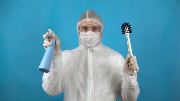 Un joven con un traje protector con un cepillo de inodoro y un dispensador en las manos. Protección contra brotes de virus. En traje antibacteriano sobre fondo azul . — Vídeos de Stock