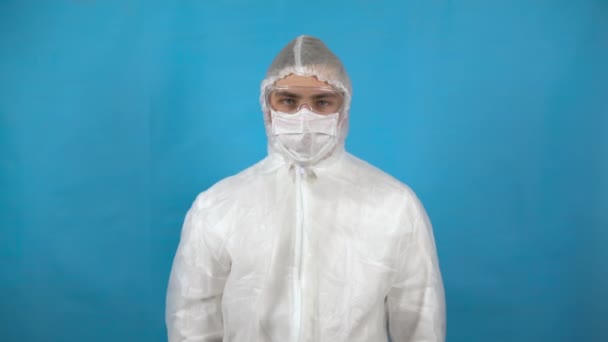 Νεαρός άνδρας σε ένα προστατευτικό δοχείο κοστουμιών στο χέρι. Ένας άνθρωπος ψεκάζει ένα αντισηπτικό σε μπλε φόντο. Προστασία από την εμφάνιση ιών. — Αρχείο Βίντεο