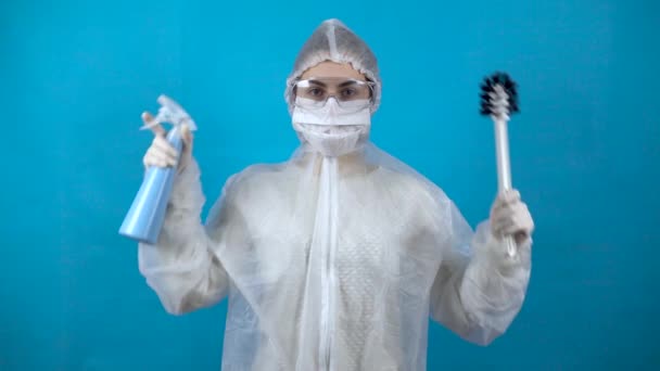 Een jonge vrouw in een beschermend pak met een toiletborstel en een dispenser in haar handen. Bescherming tegen virusuitbraken. In een antibacteriële pak op een blauwe achtergrond. — Stockvideo