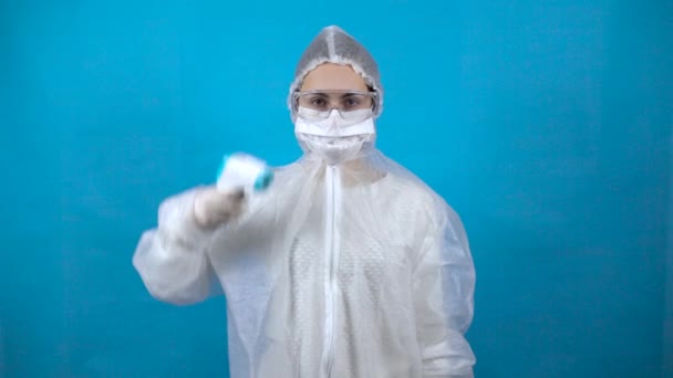 Mujer joven en traje protector con termómetro infrarrojo. La chica mide la temperatura ella misma. En un traje antibacteriano para la protección contra virus en un fondo azul . — Vídeos de Stock