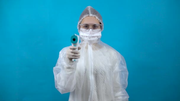 Mujer joven en traje protector con termómetro infrarrojo. La chica mide la temperatura hacia la cámara. En un traje antibacteriano para la protección contra virus en un fondo azul . — Vídeos de Stock