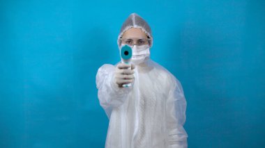 Kızıl ötesi termometresi olan koruyucu giysili genç bir kadın. Kız kameraya doğru sıcaklığı ölçüyor. Mavi arka plandaki virüslere karşı korunmak için bir anti bakteri giysisinde..