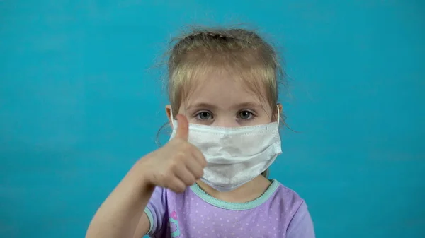 Fetiţă cu o mască medicală pe fundal albastru. Fata arată degetul mare . — Fotografie, imagine de stoc