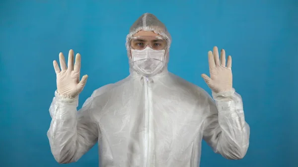 Fiatalember védőruhában. Egy férfi steril kezeket mutat kesztyűben, kék háttérrel. Vírusjárvány elleni védelem. — Stock Fotó