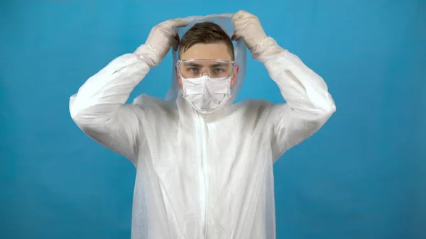 Ένας νεαρός φοράει προστατευτική στολή. Ένας άντρας φοράει μάσκα, γυαλιά και κουκούλα σε μπλε φόντο. Προστασία από την εμφάνιση ιών. — Φωτογραφία Αρχείου