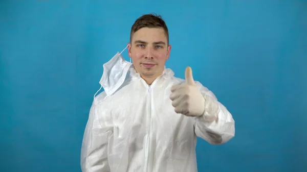 Le jeune homme enlève sa combinaison de protection et montre son pouce. Un homme se réjouit de la victoire sur l'infection sur un fond bleu. Protection contre les épidémies de virus . — Photo