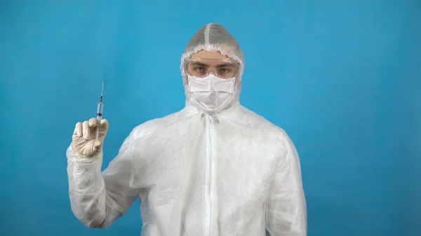 Νεαρός με προστατευτική στολή και σύριγγα στο χέρι. Ένας άνθρωπος κρατά ένα εμβόλιο κατά του ιού σε μπλε φόντο. Προστασία από την εμφάνιση ιών. — Φωτογραφία Αρχείου