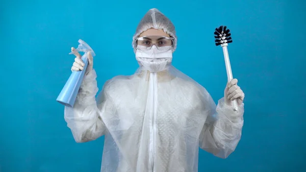Μια νεαρή γυναίκα με προστατευτική στολή με μια βούρτσα τουαλέτας και ένα δοχείο στα χέρια της. Προστασία από κρούσματα ιών. Σε ένα αντιβακτηριδιακό κοστούμι σε μπλε φόντο. — Φωτογραφία Αρχείου