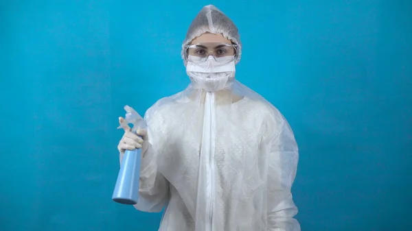 Jeune femme dans un distributeur de combinaison de protection à la main. Une fille saupoudre un antiseptique sur un fond bleu. Protection contre les épidémies de virus . — Photo