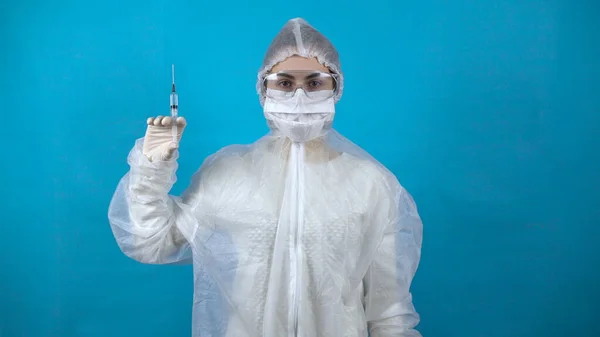 Νεαρή γυναίκα με προστατευτική στολή με μια σύριγγα στο χέρι. Ένα κορίτσι κρατά ένα εμβόλιο κατά του ιού σε μπλε φόντο. Προστασία από την εμφάνιση ιών. — Φωτογραφία Αρχείου