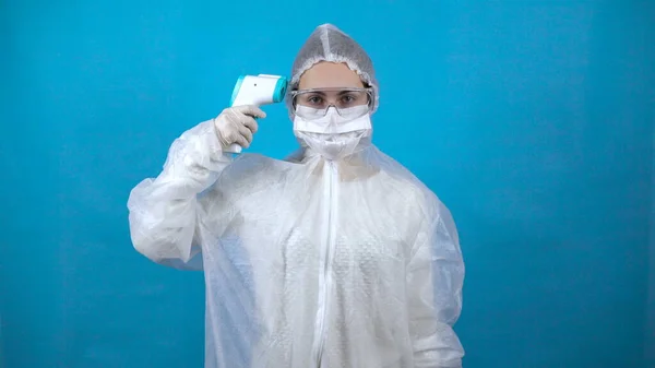 Jeune femme en combinaison de protection avec un thermomètre infrarouge. La fille mesure elle-même la température. Dans une combinaison antibactérienne pour la protection contre les virus sur un fond bleu . — Photo