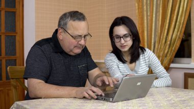 Daughter teaches father how to use a laptop. A young woman shows her old father where to click in a computer. The family is sitting in a comfortable room.
