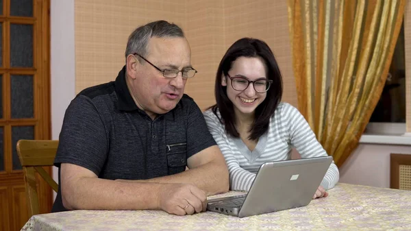A daughter and her father communicate with friends via video communication through a laptop. A young woman and an old father communicate and wave their hands while looking into a laptop. The family is — Stock Photo, Image
