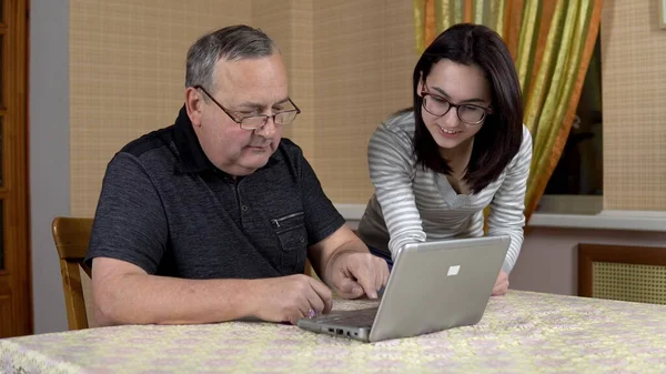 Daughter teaches father how to use a laptop. A young woman shows her old father where to click in a computer. The family is sitting in a comfortable room. A man looks at the camera and shows a thumb — Stock Photo, Image