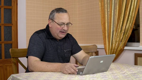 A man communicates with friends via video calling through a laptop. An old man communicates while looking at a laptop. A man in a comfortable room.