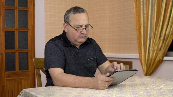 Serious man uses a tablet. Old man works with a tablet while sitting at the table. A man in a comfortable room.