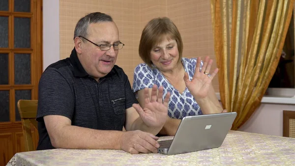 A man and a woman communicate with friends via video communication through a laptop. An old man and an old woman communicate and wave their hands while looking into a laptop. The family is sitting in — Stock Photo, Image