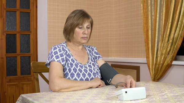 A woman uses a device to measure heart pressure. An elderly woman puts a blood pressure monitor on her hand and measures the pressure. A woman sits in a comfortable room. — Stok fotoğraf