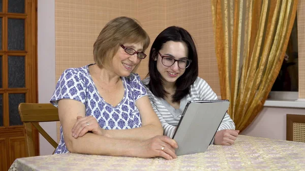 The daughter and the Mother communicate with friends via video link through the tablet. A young woman and an old mother communicate and wave their hands, looking at a tablet. The family is sitting in — Stock Photo, Image