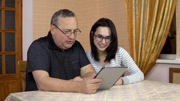 The daughter and her father communicate with friends via video link through the tablet. A young woman and an old man communicate and wave their hands, looking at a tablet. The family is sitting in a — Stock Photo, Image