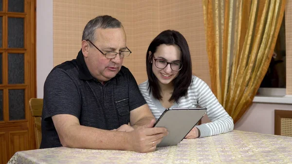 The daughter and her father communicate with friends via video link through the tablet. A young woman and an old man communicate and wave their hands, looking at a tablet. The family is sitting in a — Stock Photo, Image