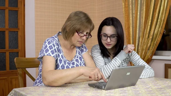 Daughter teaches mother how to use a laptop. A young woman shows her old mother where to click on a computer. The woman is surprised. The family is sitting in a comfortable room.