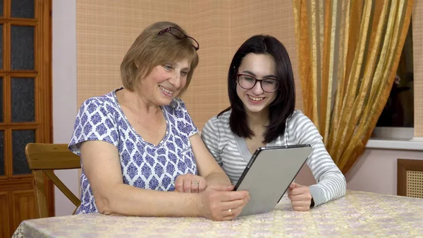 Daughter teaches mother how to use a tablet. A young woman shows her old mother where to click on a tablet. The family is sitting in a comfortable room. — Stock Photo, Image