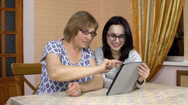 Daughter teaches mother how to use a tablet. A young woman shows her old mother where to click on a tablet. The family is sitting in a comfortable room.