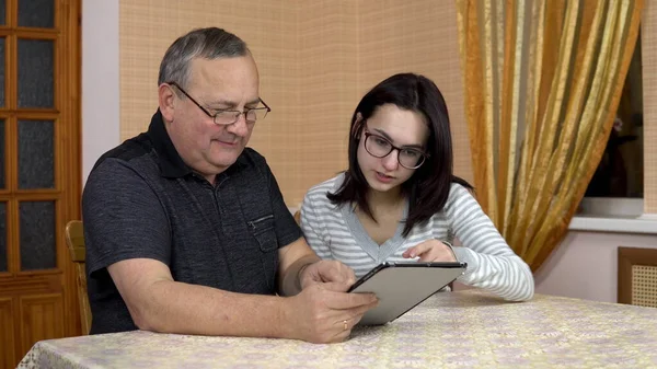 A daughter teaches her father how to use a tablet. A young woman shows her old father where to click in a tablet. The family is sitting in a comfortable room. — Stock Photo, Image