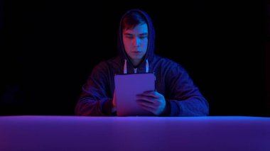 Young man in a hood with a tablet. Hacker makes a hack through the tablet. Blue and red light falls on a man on a black background.