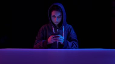 Young woman in a hood with a smartphone. Hacker makes a hack through the phone. Blue and red light falls on a woman on a black background.