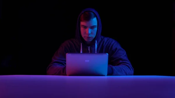 Young man in a hood with a laptop. Hacker makes a hack through a laptop. Blue and red light falls on a man on a black background. — ストック写真