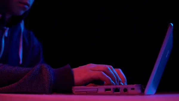 Un giovanotto col cappuccio con un primo piano portatile. L'hacker ha hackerato un portatile. Luce blu e rossa cade su un uomo su uno sfondo nero . — Foto Stock