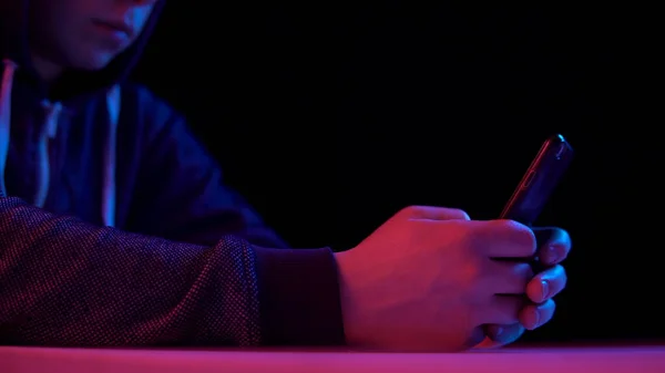 Giovane in un cappuccio con uno smartphone. L'hacker ha hackerato il telefono. Luce blu e rossa cade su un uomo su uno sfondo nero . — Foto Stock