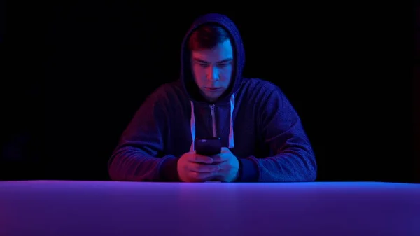 Young man in a hood with a smartphone. Hacker makes a hack through the phone. Blue and red light falls on a man on a black background. — Stockfoto